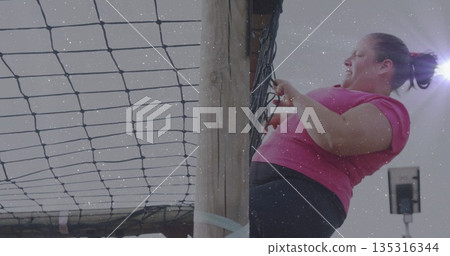 Climbing woman in pink T-shirt gripping cargo net and ascending wooden post at gym, copy space 135316344