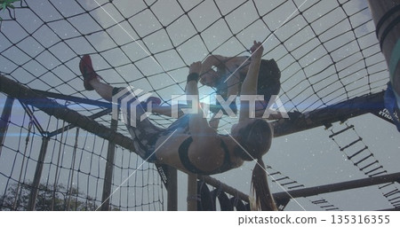 Climbing duo wearing sportswear hanging on cargo net while partner steadying at obstacle course 135316355