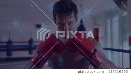 Holding red boxing gloves, shirtless boxer defending in gym ring, with ring ropes and punching bags 135316363