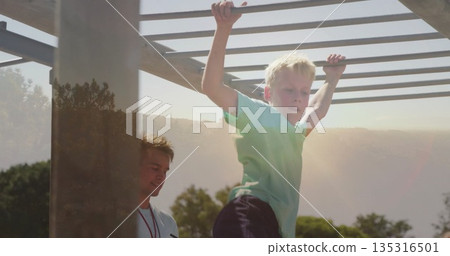 Climbing boy wearing green T-shirt on monkey bars at playground, supervisor wearing whistle lanyard Climbing boy wearing green T-shirt on monkey bars at playground, supervisor wearing whistle lanyard 135316501