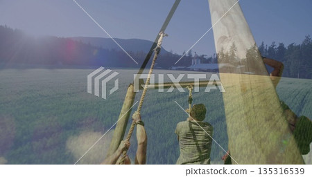 Climbing obstacle frame in green field wearing gloves and straps gripping thick ropes, barn silos 135316539