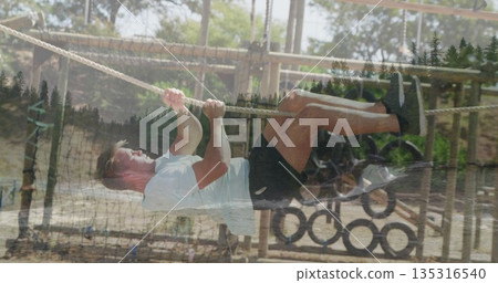 Maneuvering male fitness participant gripping rope at forest obstacle course, under pine trees Maneuvering male fitness participant gripping rope at forest obstacle course, under pine trees 135316540