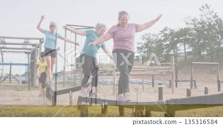 Six women in sportswear balancing on wooden beam along obstacle course, with metal scaffolding 135316584