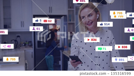 Streaming live woman wearing polka-dot blouse and earbuds holding smartphone in kitchen, copy space 135316599