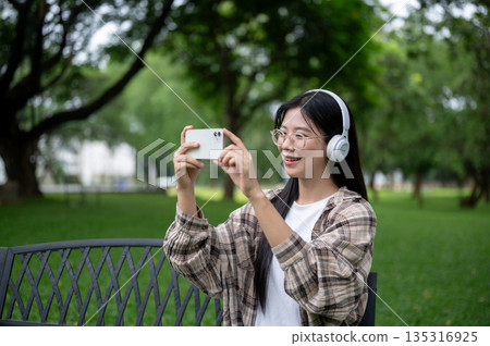 Young asian glasses woman wearings headphone taking pictures on phone while sitting on bench in park Young asian glasses woman wearings headphone taking pictures on phone while sitting on bench in park 135316925