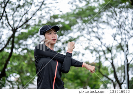 Young asian woman in sportswear wears headphones arms stretching before exercise or workout in park 135316933