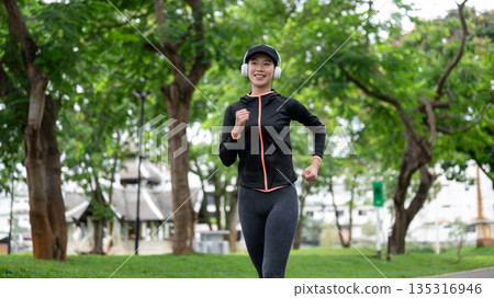 Young asian woman in sportswear with headphone on listening to music as exercise by running in park. Young asian woman in sportswear with headphone on listening to music as exercise by running in park. 135316946