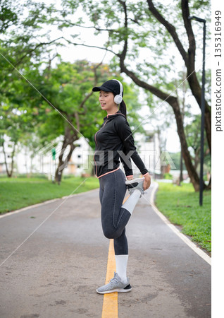 Young asian woman in sportswear and headphone stretching her legs before exercise or workout in park 135316949
