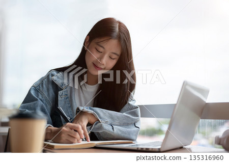 Young asian woman in denim jacket writing in notebook with pen aside laptop on wooden table in cafe. 135316960