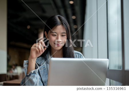 Young asian woman in denim jacket holding pen and smiling as she looking at laptop on table in cafe 135316961