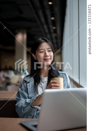 Smiling young asian woman holding coffee while sitting with laptop on table in the cafe or lounge. Smiling young asian woman holding coffee while sitting with laptop on table in the cafe or lounge. 135316971