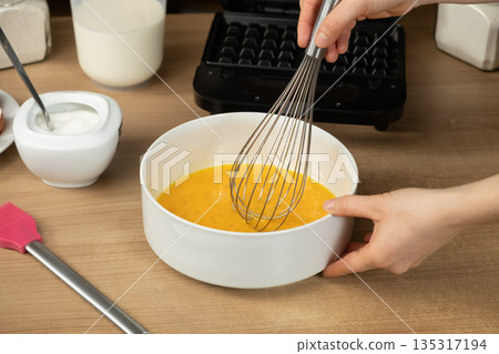 Hand Using a Whisk to Mix Raw Eggs in a White Bowl for Baking Waffles Hand Using a Whisk to Mix Raw Eggs in a White Bowl for Baking Waffles 135317194