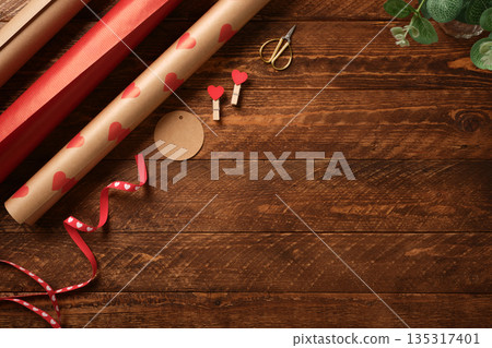 Gift wrapping paper with red hearts, ribbon, and scissors on wooden background. Valentine's Day or wedding gift preparation. Copy space Gift wrapping paper with red hearts, ribbon, and scissors on wooden background. Valentine's Day or wedding gift preparation. Copy space 135317401