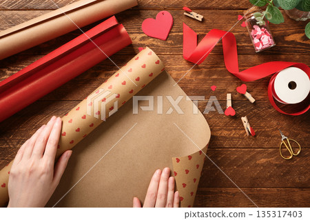 Hands unrolling heart-patterned craft paper on wooden table. Gift wrapping for Valentine's Day or wedding with red ribbon, scissors, and decor. 135317403