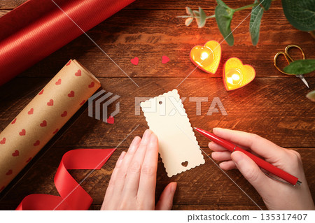 Romantic flat lay with hands writing gift tag on wooden table. Heart candles, red ribbon, wrapping paper. Handmade greeting with copy space 135317407