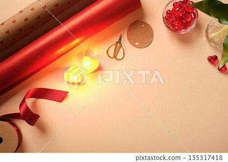 Flat lay of Valentine's Day gift wrapping supplies on a beige background. Red paper, heart candles, ribbon, and scissors with copy space. Flat lay of Valentine's Day gift wrapping supplies on a beige background. Red paper, heart candles, ribbon, and scissors with copy space. 135317418