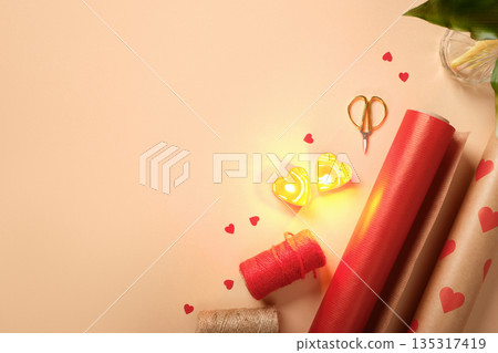 Romantic gift wrapping supplies on beige background. Red paper, heart candles, ribbon, and scissors with copy space. Romantic gift wrapping supplies on beige background. Red paper, heart candles, ribbon, and scissors with copy space. 135317419