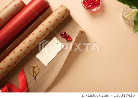 Gift wrapping supplies with red paper rolls, ribbon, scissors, tag and heart decorations on beige background. Copyspace. Gift wrapping supplies with red paper rolls, ribbon, scissors, tag and heart decorations on beige background. Copyspace. 135317420