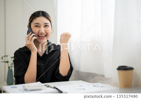 Smiling asian businesswoman or office worker making a fist while talking on phone at working table. Smiling asian businesswoman or office worker making a fist while talking on phone at working table. 135317490