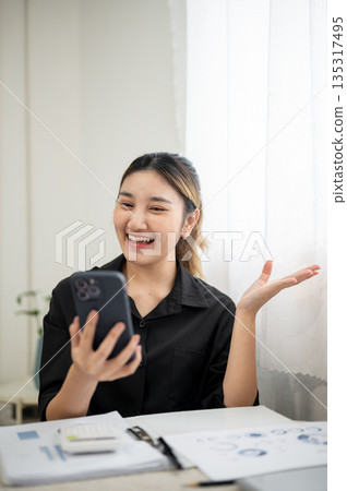 Asian businesswoman or office worker opening palm looking happy during phone call at working table. Asian businesswoman or office worker opening palm looking happy during phone call at working table. 135317495