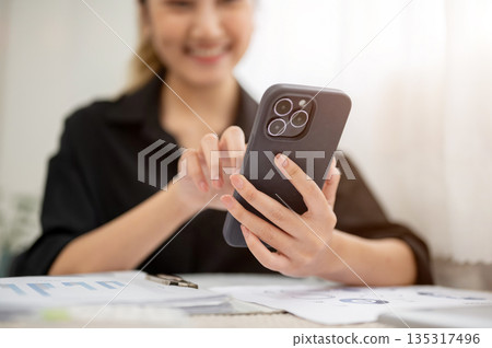 Close up of asian businesswoman or office worker smiling while looking at phone over working table. Close up of asian businesswoman or office worker smiling while looking at phone over working table. 135317496