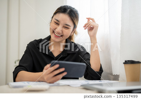 Smiling asian businesswoman or office worker looking at phone holding horizontally on table. 135317499