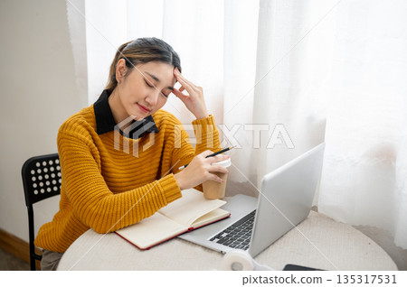 Tired asian woman or student office worker holding pencil and coffee closing eyes in front of laptop Tired asian woman or student office worker holding pencil and coffee closing eyes in front of laptop 135317531