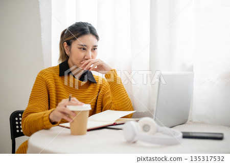 Serious asian woman student office worker holding pencil and coffee while looking at laptop on table Serious asian woman student office worker holding pencil and coffee while looking at laptop on table 135317532