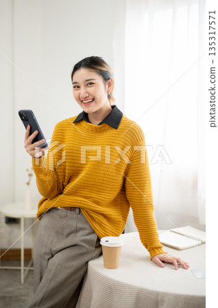 Happy young asian woman holding and look away from phone while standing leaning against white table. Happy young asian woman holding and look away from phone while standing leaning against white table. 135317571