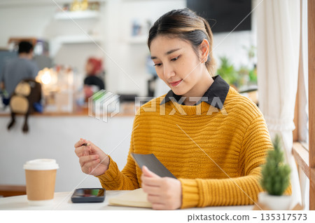 Pretty asian woman holding pencil and looking at notebook aside phone while sitting at cafe's table. Pretty asian woman holding pencil and looking at notebook aside phone while sitting at cafe's table. 135317573