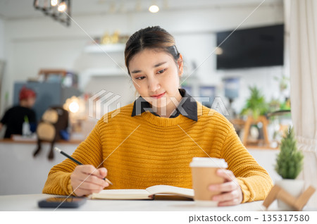 Asian woman holding pencil over notebook and looking at coffee cup while sitting at cafe's table. Asian woman holding pencil over notebook and looking at coffee cup while sitting at cafe's table. 135317580