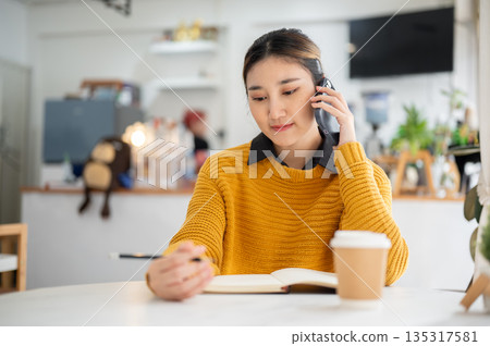 Young asian woman holding pencil over notebook and making a phone call while sitting at cafe's table Young asian woman holding pencil over notebook and making a phone call while sitting at cafe's table 135317581