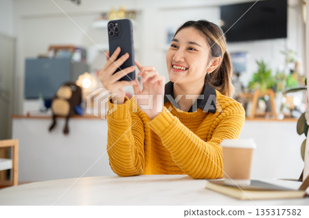 Smiling asian woman holding and looking at phone typing or taking photos while sitting at cafe table Smiling asian woman holding and looking at phone typing or taking photos while sitting at cafe table 135317582