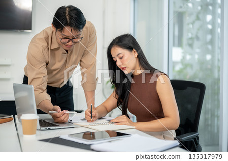 Businessman or office worker talking and looking at notebook explaining to a woman at working table. Businessman or office worker talking and looking at notebook explaining to a woman at working table. 135317979