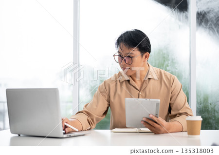 Asian businessman or office worker holding tablet and using a laptop while sitting at working table. 135318003
