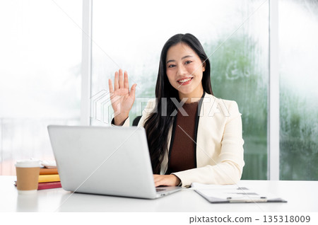 Asian businesswoman or office worker waving a hand as sitting in front of laptop at working table. Asian businesswoman or office worker waving a hand as sitting in front of laptop at working table. 135318009