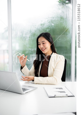 Asian businesswoman or office worker in suit holding pen explaining to a laptop at working table. Asian businesswoman or office worker in suit holding pen explaining to a laptop at working table. 135318012