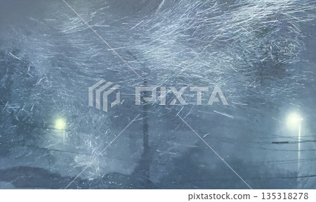 An abstract, cinematic view of a ferociously blowing blizzard at night, with swirling snow streaks caught in the bright glow of a street lamp against a dark, hazy winter background. 135318278