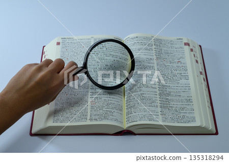 A person looking up a dictionary with a magnifying glass 135318294