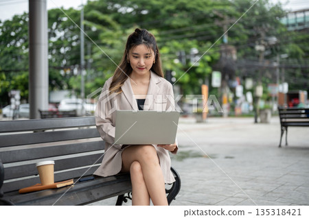 Asian woman using or working on laptop aside book and coffee while sitting on bench in park. 135318421