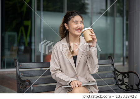 Smiling asian woman drinking coffee while sitting with laptop on metal bench in front of the store. Smiling asian woman drinking coffee while sitting with laptop on metal bench in front of the store. 135318428