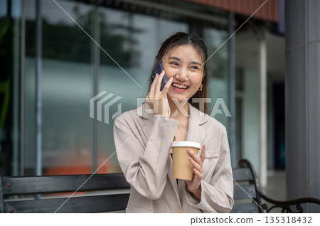 Smiling asian woman holding coffee during phone call while sitting on metal bench in front of store. 135318432