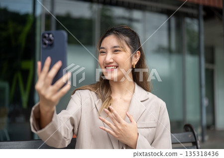 Smiling asian woman holding phone taking picture or video call while sits on bench in front of store Smiling asian woman holding phone taking picture or video call while sits on bench in front of store 135318436