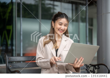 Smiling asian woman holding laptop typing or watching video while sitting on bench in front of store Smiling asian woman holding laptop typing or watching video while sitting on bench in front of store 135318441