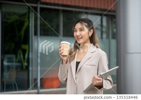 Beautiful asian woman holding coffee cup and carrying a laptop while standing in front of the shop. Beautiful asian woman holding coffee cup and carrying a laptop while standing in front of the shop. 135318446