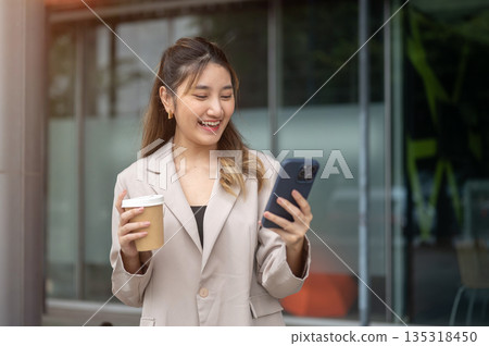 Asian woman holding coffee and excitedly looking at phone while standing in front of a cafe or store Asian woman holding coffee and excitedly looking at phone while standing in front of a cafe or store 135318450