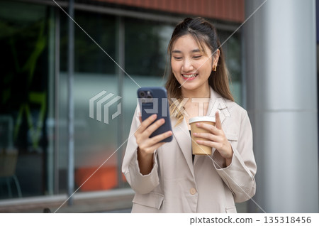 Smiling asian woman holding coffee and looking at phone while standing in front of a cafe or store. Smiling asian woman holding coffee and looking at phone while standing in front of a cafe or store. 135318456
