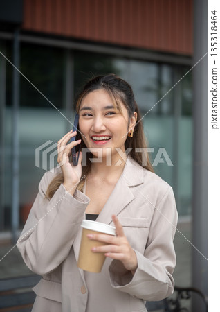 Pretty asian woman holding coffee during a phone call while standing or walking in front of a cafe. Pretty asian woman holding coffee during a phone call while standing or walking in front of a cafe. 135318464