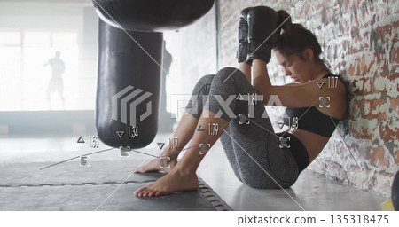 Sitting athlete wearing boxing gloves, leggings by brick wall on gym floor, with heavy punching bag Sitting athlete wearing boxing gloves, leggings by brick wall on gym floor, with heavy punching bag 135318475