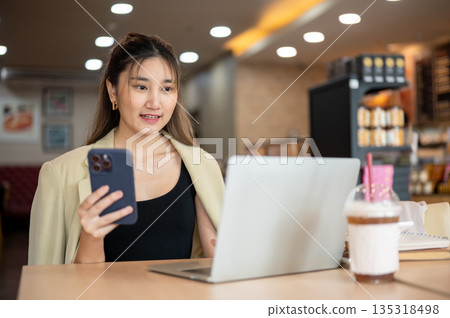 Smiling asian woman holding phone and looking at laptop while sitting at wooden table in the cafe. 135318498